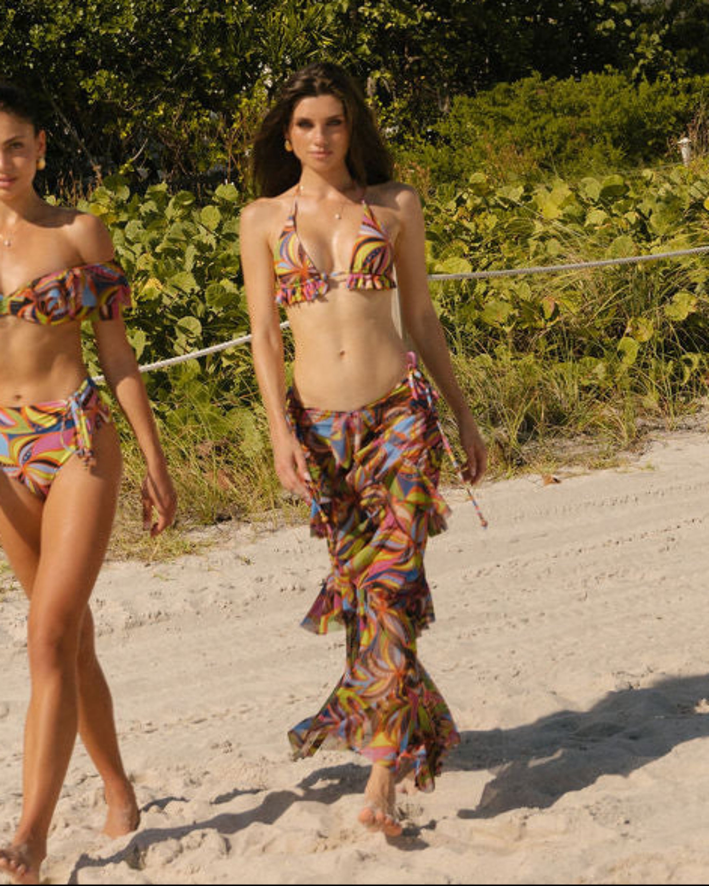 Two women in TRUSSO SWIM walking on a sandy beach with bold printed swimwear and a coverup skirt